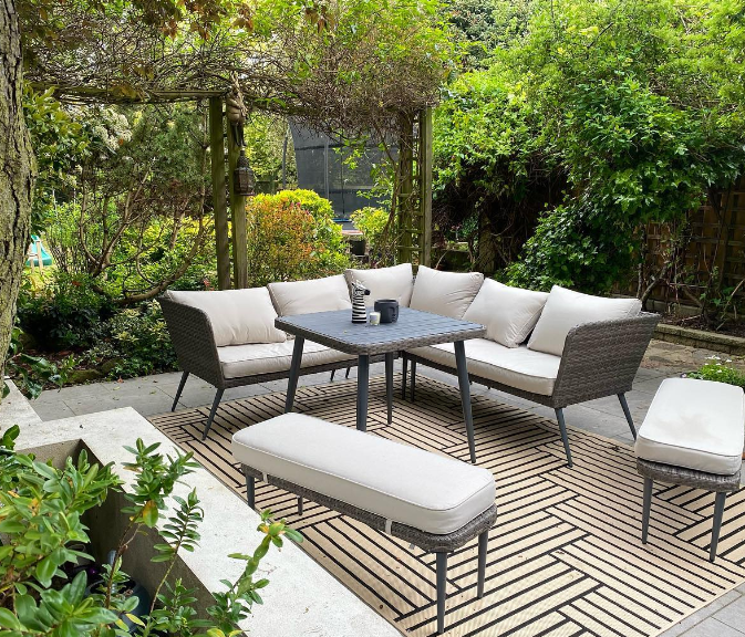 modern outdoor garden sofa in brown rattan with table and two benches, on patterned outdoor rug, with tall greenery and trellis surrounding it