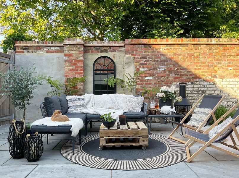 picturesque walled garden in exposed brock, with outdoor garden mirror, round outdoor rug and grey rattan and metal corner garden sofa and chaise, with two grey deckchairs, chimenia and reclaimed pallet coffee table.