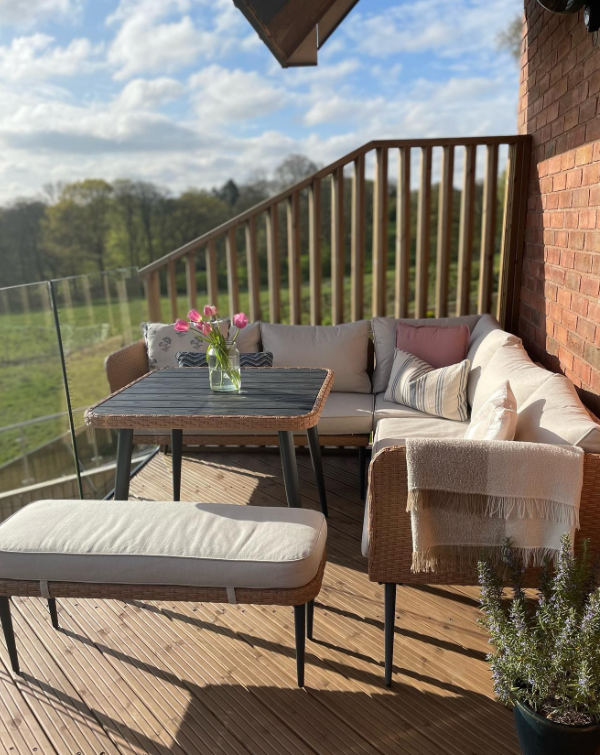 brown beige rattan outdoor corner sofa set with bench and table in the corenr of a modern apartment balcony, with potten plants and field with blue sky beyond.