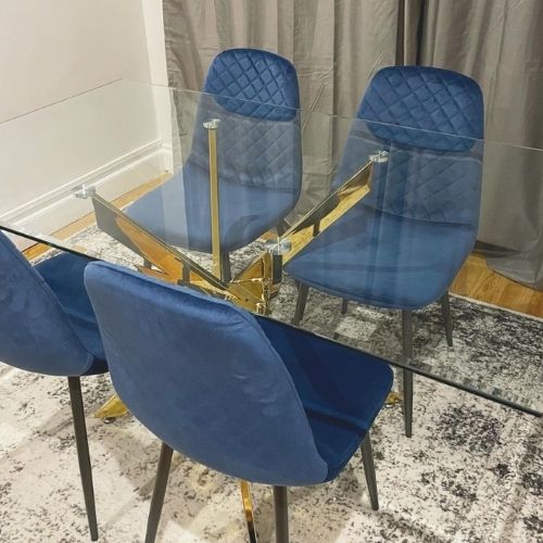 rectangular glass dining table with gold nested starburst legs, 4 navy velvet dining chairs with black legs, on silver and dark grey abstract patterned rug, on a warm yellowish wood floor with white walls and grey curtains.