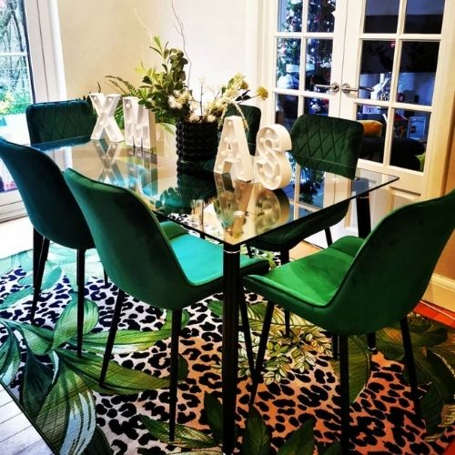modern maximalist dining area featuring glass and black metal dining table and 6 green velvet chairs, with green vase and green/white flowers. Sat on a leopard print rug that also has large leaves printed on it. Walls are white. 