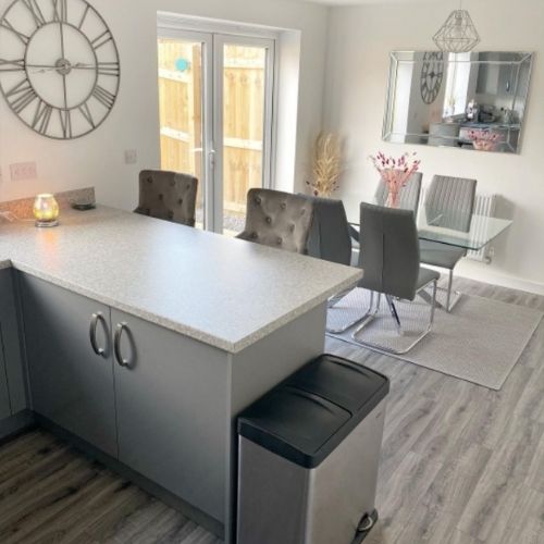 Open plan kitchen-diner space with glass and chrome table with silver nested starburst legs and 4 grey faux leather cantilever chairs on a pale grey rug, at the rear of the room. Kitchen bar table and barstools in foreground.