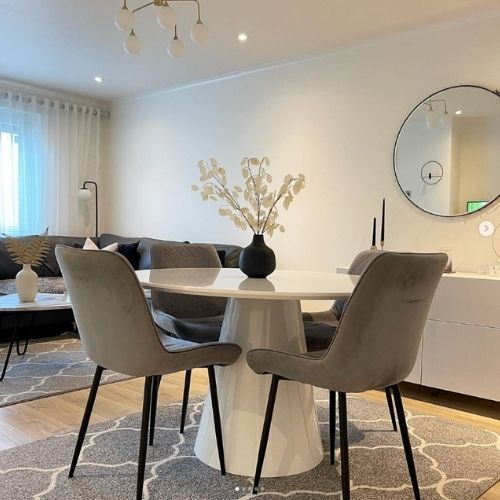 white gloss round dining table with pedestal plinth and 4 grey velvet chairs on a grey rug with cream moroccan trellis pattern. In background, sofa area can be seen with matching rug under coffee table. Round wall mirror on wall above white storage units.