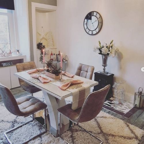 bright dining area with ash wood effect flooring, cream-white walls, large wall clock, white gloss dining table with X shaped legs and chrome elements, 4 faux leather cantilever chairs dressed with table runner and pink candles. 