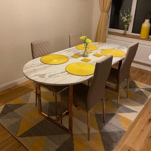 Marble effect and gold dining table with 4 tall backed faux leather dining chairs, with yellow place mats, curtains and accessories, on a yellow grey and white rug in triangular patterns.