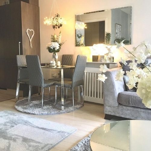 Rectangular glass and chrome dining table with 4 grey faux leather dining chairs, on round grey rug, beside a sofa and round coffee table, with rectangular grey rug. Lamps and white flowers decorate the room.