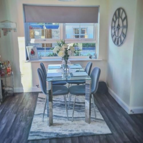 glass and chrome dining table on white and grey rug, on darker ash wood floor, with white walls. Also 4 upholstered grey velvet chairs with silver legs