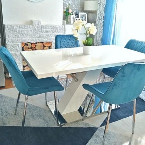 bright dining room with white walls and cyan blue curtains, with white high gloss and chrome dining table and 4 blue velvet chairs on blue and grey patterned rug.