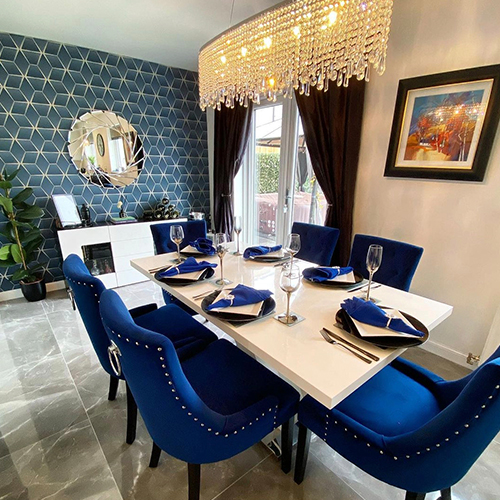 dining room wallpaper ideas - bright dining area with white gloos table and blue velvet chairs, marble effect tile flooring, navy wallpaper to one wall with white geometric pattern.