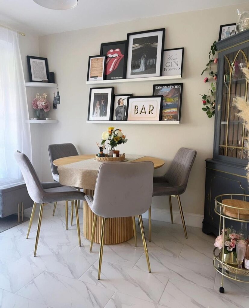 modern eclectic dining space in open plan kitchen - gallery wall of fun artwork on wall, round wooden pedestal table, grey velvet chairs with gold legs, naby blue dresser and gold drinks trolley