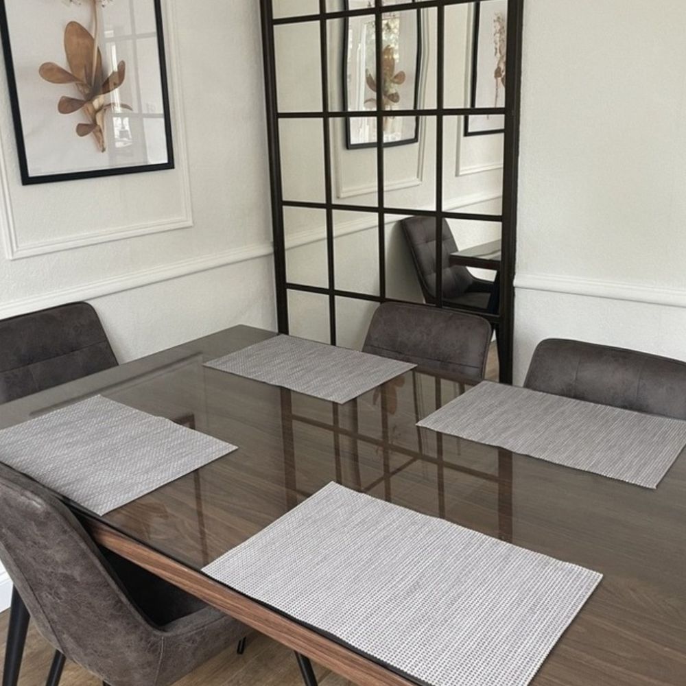 wooden dining table with grey chairs, with glass table top protector on top. 4 grey place mats. Large wall window style mirror. 