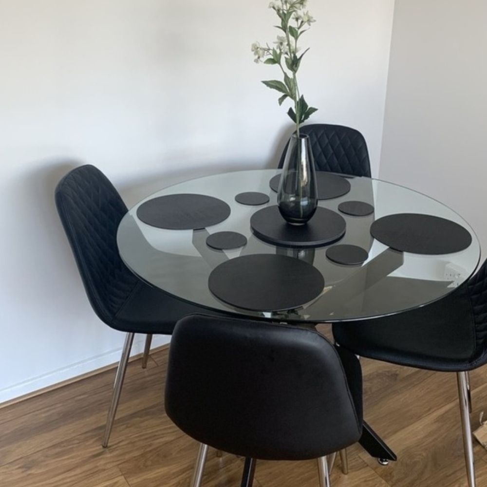 round glass dining table with black metal starburst legs, and 4 black faux leather dining chairs with silver tapered legs in a white room