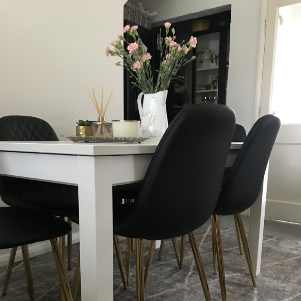 white rectangular dining table with 4 black faux leather dining chairs with gold tapered legs. Gold and white accessories on the table including candle jars, white jug with pink flowers and gold plant pot. 