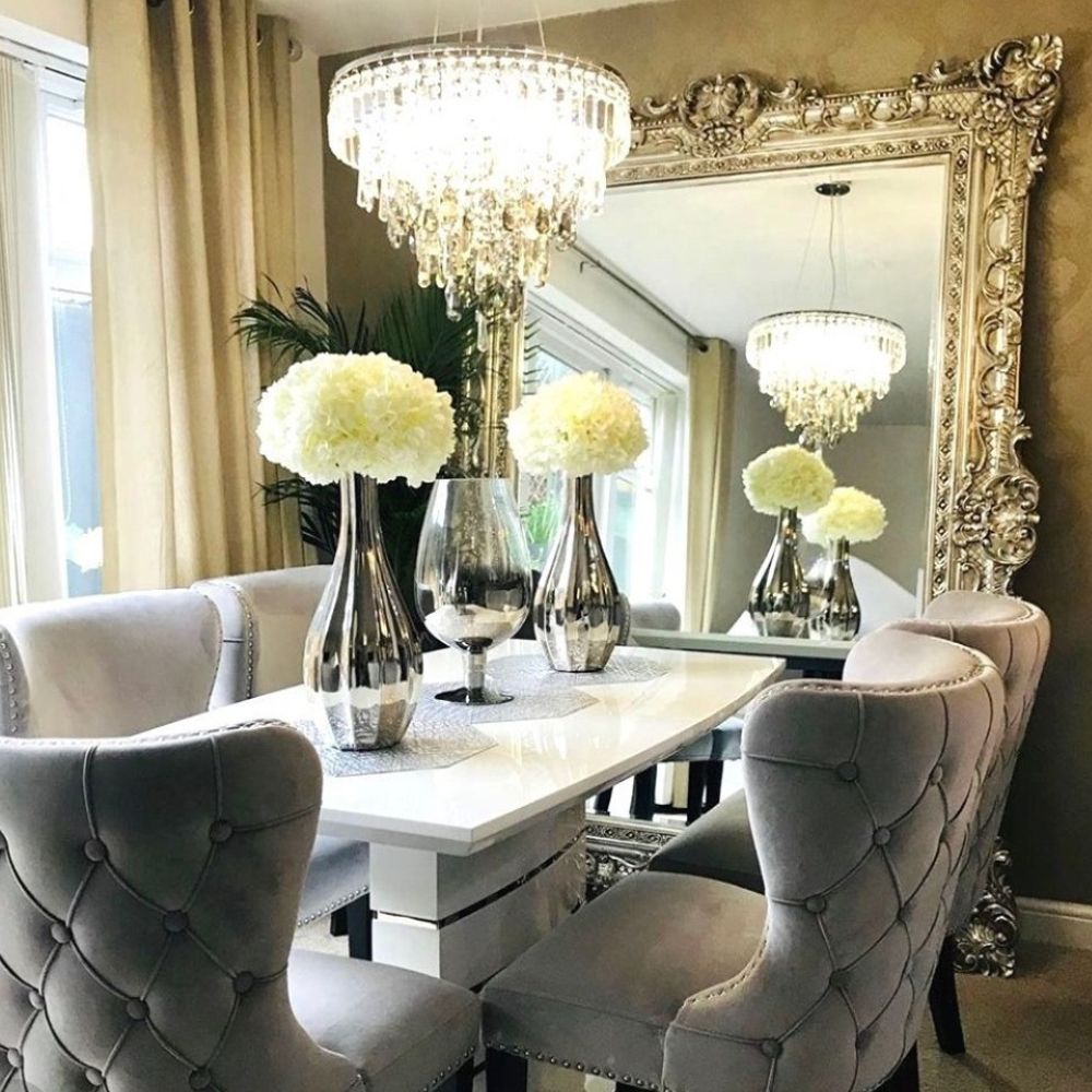 Elegant dining room with large gold gilt frame wall mirror leaning on back wall, white high gloss extending dining table with 5 grey velvet chairs, 2 tall vases with cream roses, modern glass crystal chandelier. 