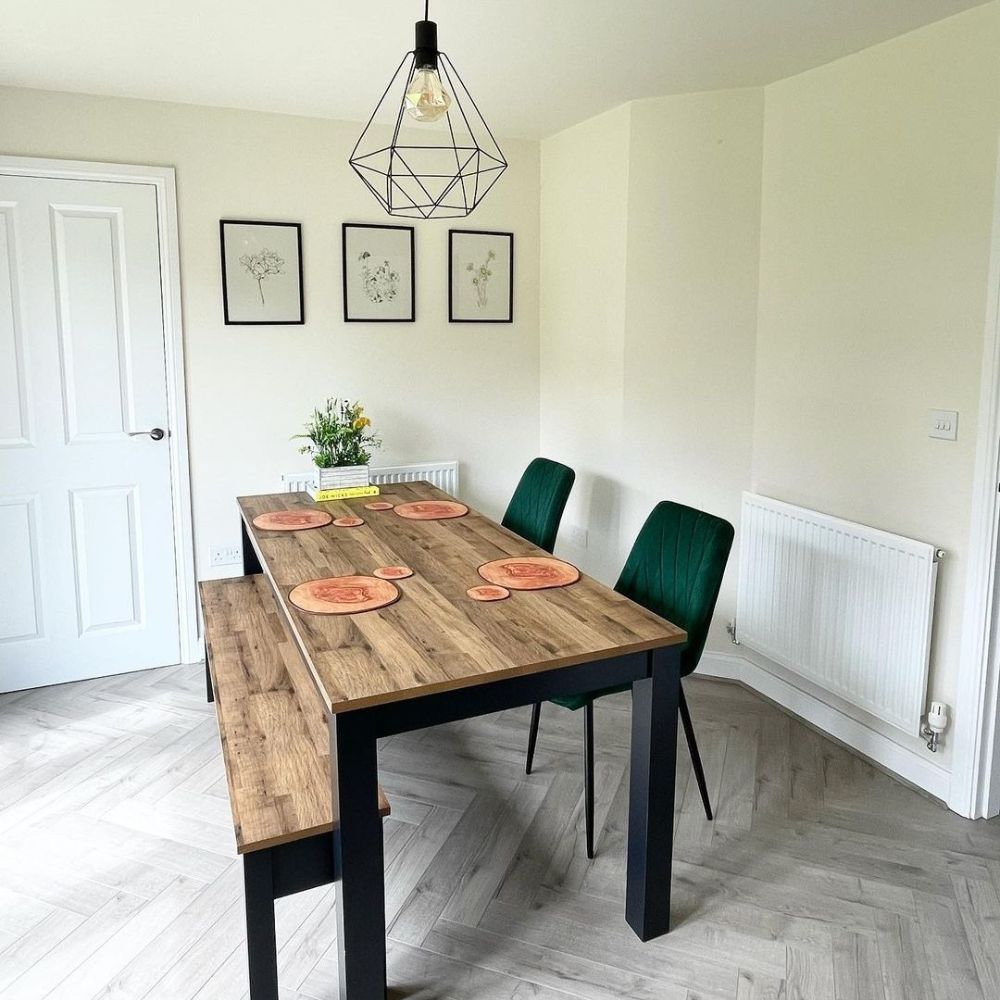 rectangular wood table with black metal legs, wooden bench and  green velvet chairs with black legs on pale ash floor with cream walls, 3 photo frames on wall, black industrial wire frame light fixing