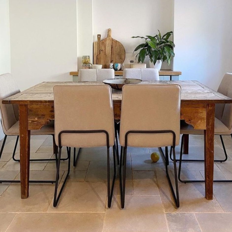 rustic wooden farmhouse table with 6 fabric chairs, all with black industrial style wire frame legs, on stone tiles, with white walls. Wooden chopping boards and pot plant visible in background. 