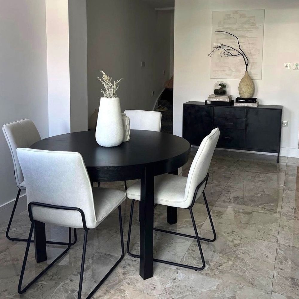 round black wooden table with 4 modern dining chairs. Chairs are cream fabric, with black metal wire-frame legs on grey marble floor