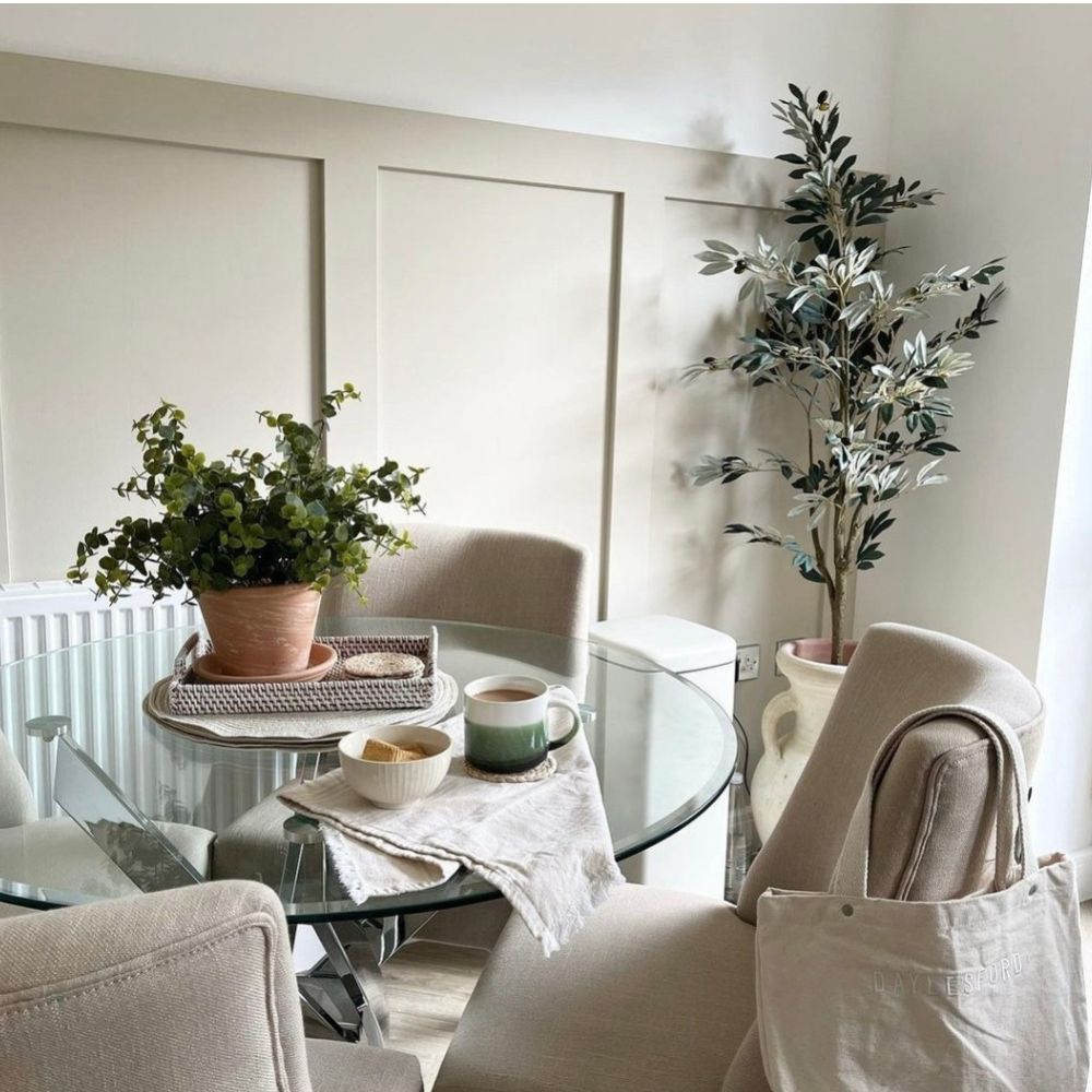 Neutral elegant dining room with beige wall, round glass dining table, 4 beige fabric chairs, soft linens, stoneware cups, terracotta pot with green plant, tall urn in corner with eucalyptus leaves