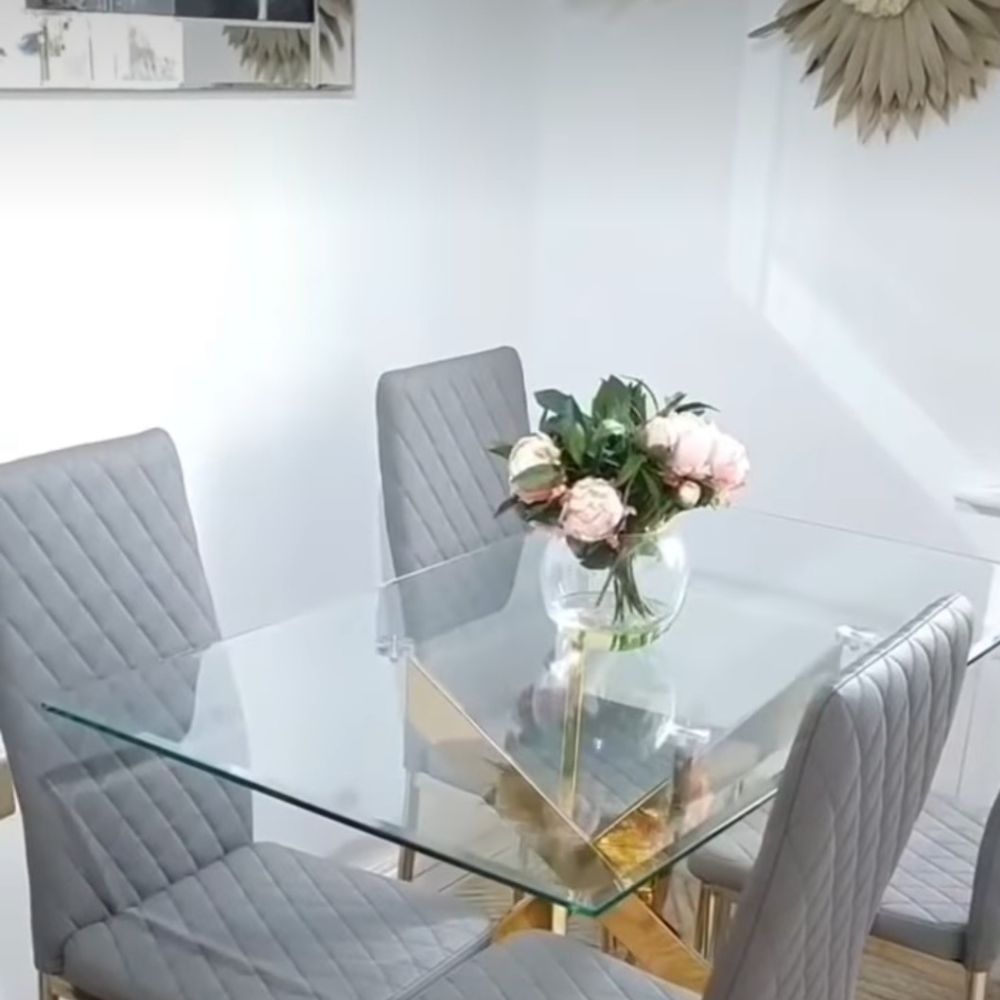 Grey dining room with rectangular glass dining table that has gold meta starburst legs. In corner of room, with white walls, wooden floors, glass bowl with pink peonies on tabletop. Large wall mirror and gold accessories on wall.