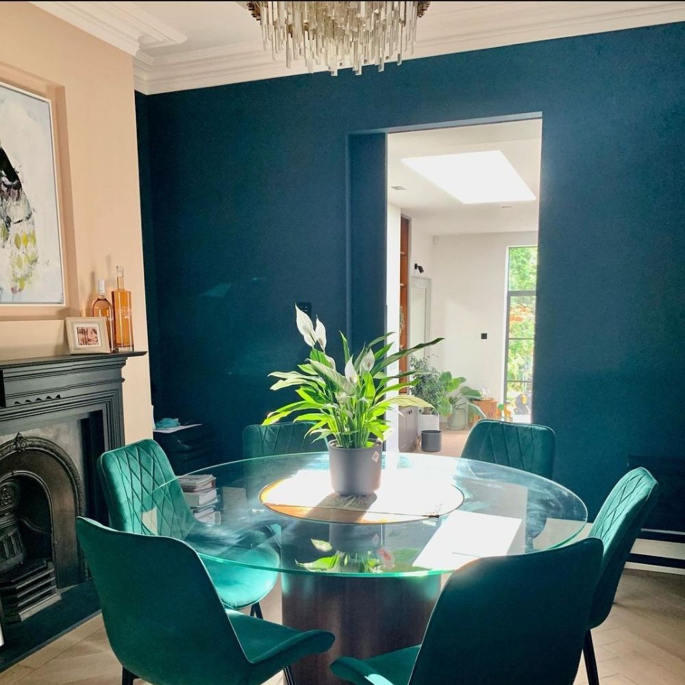 round glass dining table with black pedestal leg, surrounded by 6 green velvet dining chairs with black legs, in dining room that has feature black period fireplace, one forest green feature wall and modern chandelier light. 