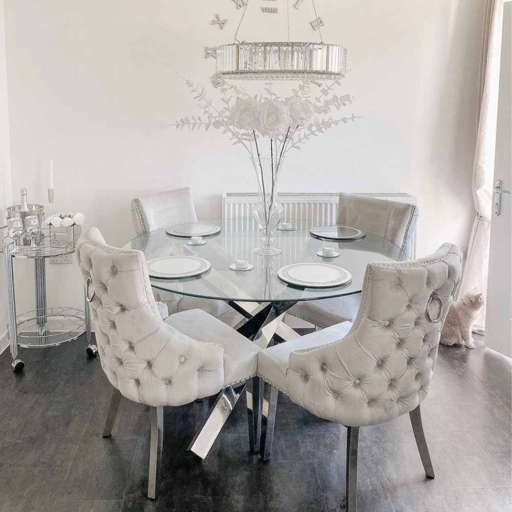 elegant dining room featuring grey floor, round glass dining table with chrome starburst legs, 4 velvet tall-back knockerback chairs, chrome drinks trolley, glass vase with white flowers, hanging glass and chrome light fixture