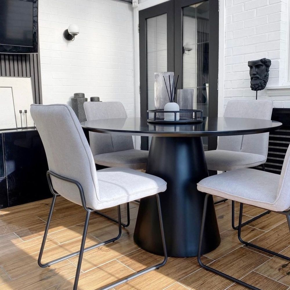 round black semi-gloss dining table with 4 Industrial style fabric dining chairs that have black metal wire-frame legs. in back and white room with warm wooden flooring