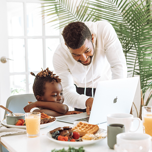 A young child with dark skin and natural hair is sat on a chair at a small dining table covered in breakfast items, looking at a laptop place amongst food/crockery. An adult man with dark skin leaning over and pointing at the laptop screen. Both are smiling. 