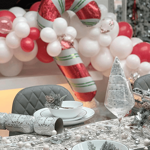 animated GIF showing 2 images of hte same room - close up and wide shot - featuring modern glass and chrome table and bench seat with grey velvet seat, and red and white christmas themed balloon arch. 