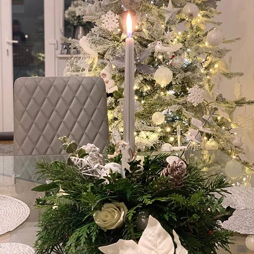 clsoe up of silver taer candle in christmas wreath on a glass table. A white and gold christmas tree is visible in the background. 