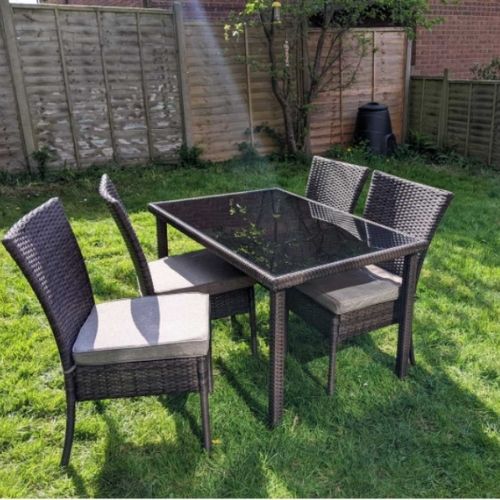 garden furniture - brown rattan chairs with grey cushions and brown rattan dining table with black glass top place on lawn in garden with fence and trees in background 