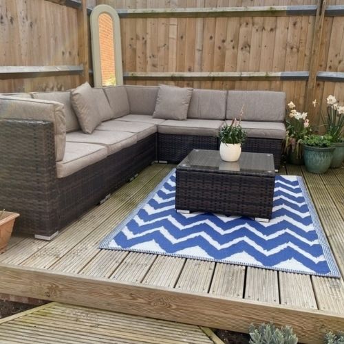 brown rattan modular outdoor furniture sofa placed on wooden deck with blue and white outdor rug, rattan coffee table, and deocrated with miroors and plant pots