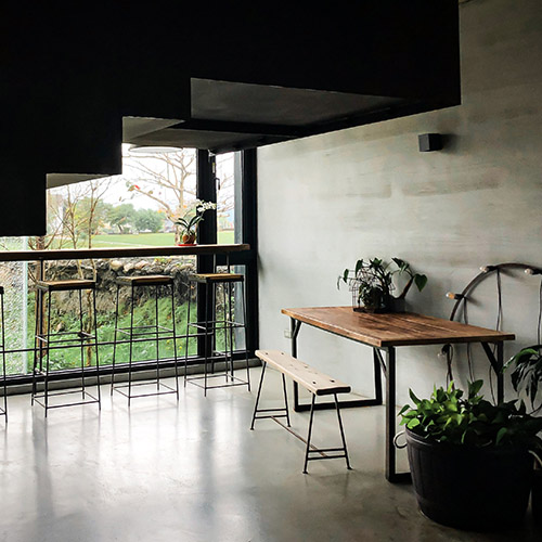 Industrial style dining area wth large wood table with black metal legs and bench with hairpin legs. Wooden bar and tall wooden barstools infront of large window. Grey stone wall with black accents and green potted plants in black tubs.