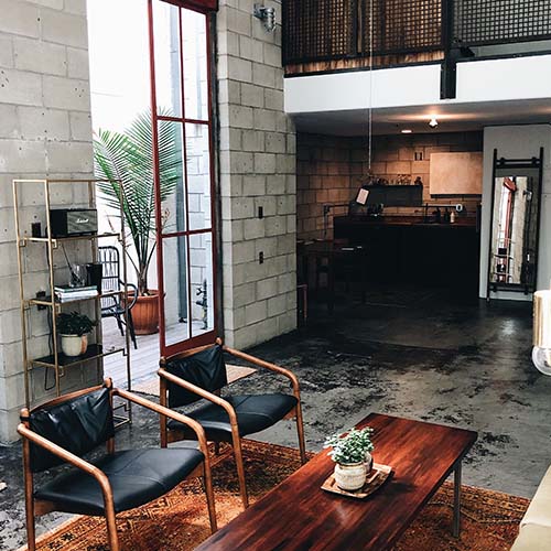 Industrial style open plan living room and kitchen area featuring eposed grey block briks, warm dark wood table and wood frame chairs with black leather seats. Low hanging lights, gold shelving and warm, worn rug under table. 