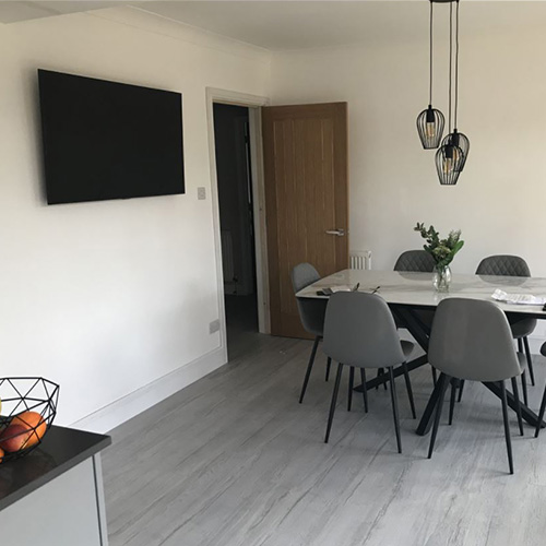 Bright grey and white open plan kitchen-diner area with marble-effect dining table with black starburst legs, and 6 grey faux leatehr chairs with tapered black legs.