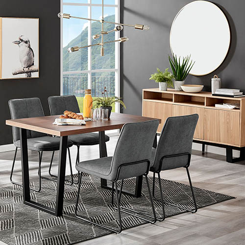 another industrial dining table with thick wood effect tabletop and chunky black metal legs, with 4 grey fabric upholstered chairs on black hairpin metal legs, with gold pendent hanging light fixings. Wood and black metal sideboard in background with round gold framed wall mirror.