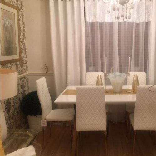 white high gloss dining table with trestle table inspiration, with gold geometric frame legs, and  6 white faux leather dining chairs with gold legs, in dining room with wood floor and gold baroque scroll pattern wallpaper, candlesticks and chandelier style light fixing. 