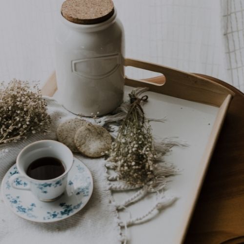 shabby chic accessories image features wooden tray with ceramic jar and cork lid, dried flowers, oat biscuits, china tea cup and saucer on white linen sheet.