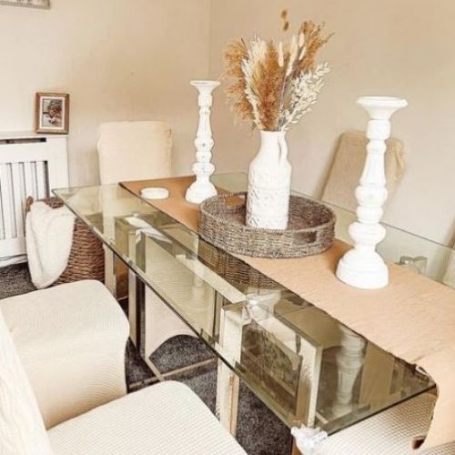 glass rectangular dining table with gold nested starburst legs, and 6 high back dining chairs in oatmeal coloured fabric chair covers. 2 white pillar candlesticks, a wicker basket with ceramic jug vas and dried flower arrangement on table with hessian table runner. 
