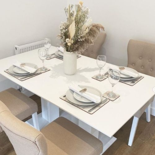 white gloss dining table with 2 pillar legs and 4 beige upholstered fabric chairs, with grey table ware, ceramic vase with drief flowers
