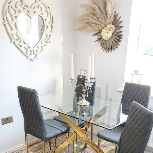 glass rectangular dining table with gold nested starburst legs and 4 grey faux leather dining chairs with shabby chic candlestick on table, heart-shaped mirror on wall in scrolled frame and pampas grass