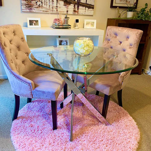 round glass dining table with nested starbusrt chrome legs and 2 pink-grey velvet knockerback chairs n a round pink rug.