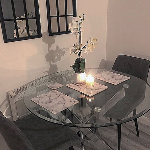 round glass dining table with chrome silver nested legs and 2 grey velvet chairs. White marble effect placemats and candle. 2 black framed window-style mirrors on the wall.