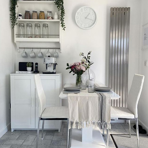 Bright kitchen area with white cabinet, coffeemachine and mugs and shelves with jars/cups. Grey tiled floor. White high gloss dining table with 2 column plinth legs, 2 white faix leather chairs. Table is dressed with linen white and black tablecloth and flower arrangement.