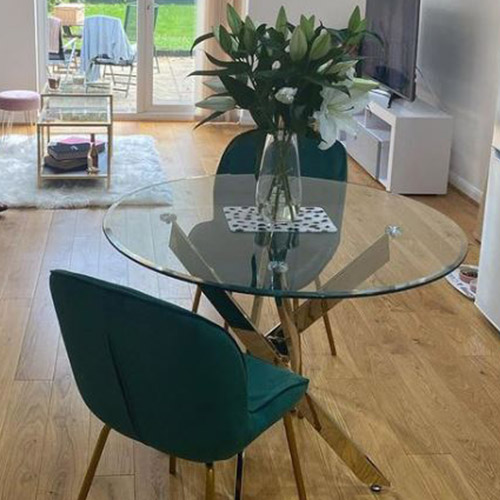 round glass dining table with nested gold shiny legs and 2 green velvet chairs with gold legs.