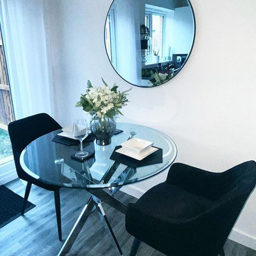 round dining table with 2 black velvet chairs, with black hexagonal placemats and white square crockery. Wine glasses and vase of white flowers. 