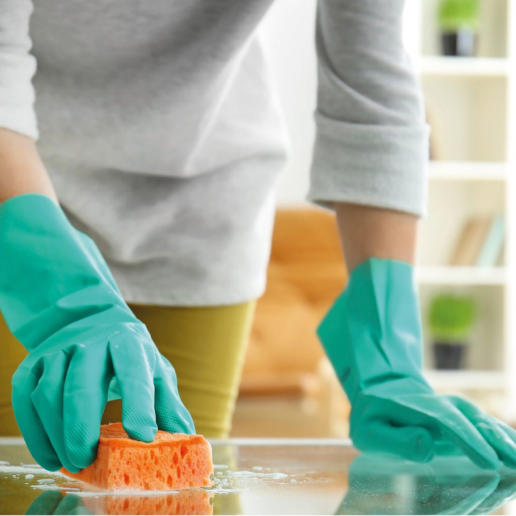 Image shows a person wearing green rubber gloves cleaning a table with an orange sponge