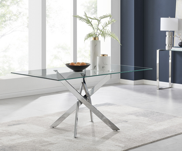 Image shows a dining table with glass top and chrome legs in a criss-cross pattern, set in a room with large windows and a dark blue wall.