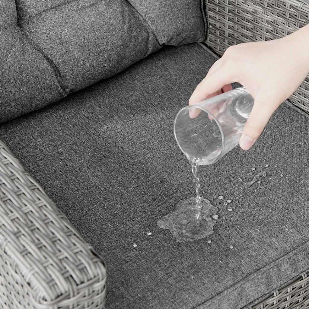 close up of outdoor rattan furniture and grey cushion - a hand is pouring a glass of water into the cushion to demostrate its waterproofing.