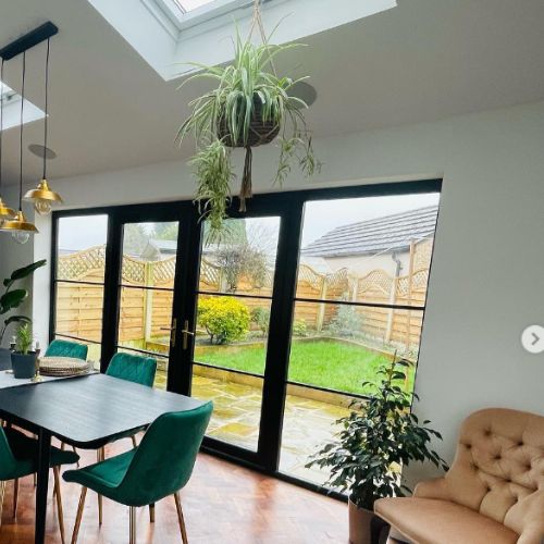 dining room space with large black framed french windows, black dining table and green velvet dining chairs, with decorative house plants.