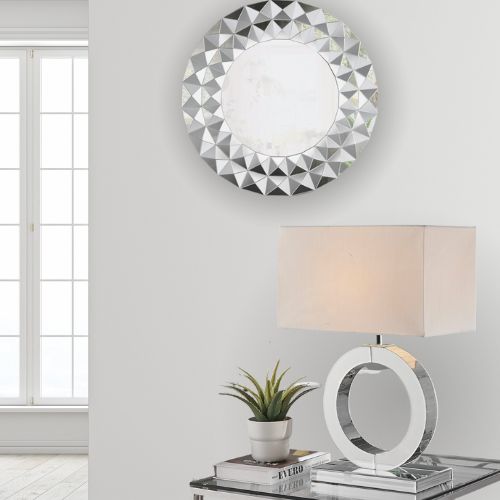 white room with chrome side table, large statement table lamp that has a mirrored stand and halo design and cream rectangular lampshade, and a round wall mirror with cut diamond pattern.
